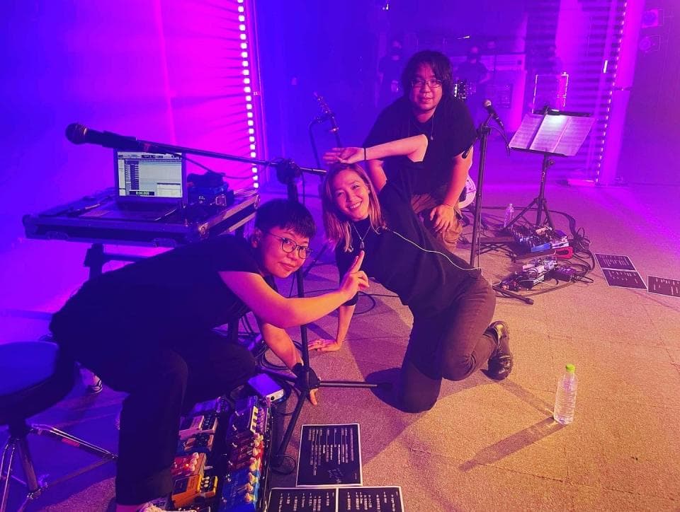 這張照片是三個朋友在舞台上表演,其中一個女孩正跪在地上唱歌,另一個女孩則坐在她旁邊伴奏。