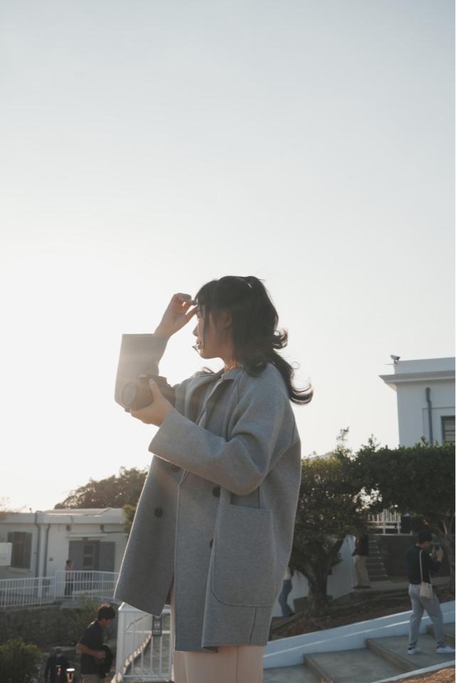 這張照片是一個穿著灰色外套的女孩,她站在陽光下,手裡拿著相機,正準備拍攝照片。