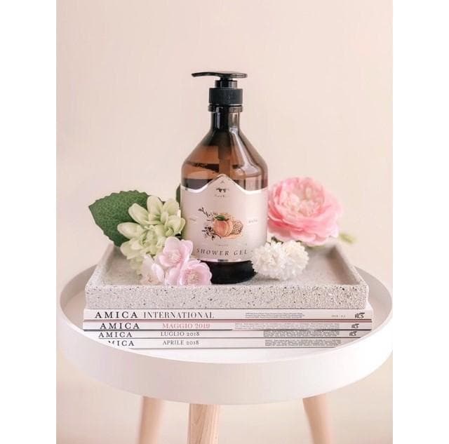這張照片是一瓶棕色玻璃瓶的沐浴乳,上面貼著一個桃色的標籤,寫著「SHOWER GEL」,瓶子上還印著一個桃子的圖案,瓶子放在一個水泥色的託盤上,託盤上還擺著一些粉紅色的花。