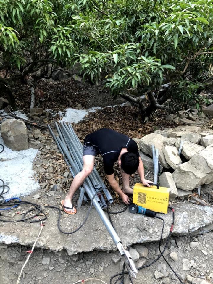 這張照片是一個男人正在用電焊機焊接金屬管,他正在一片樹林中工作。