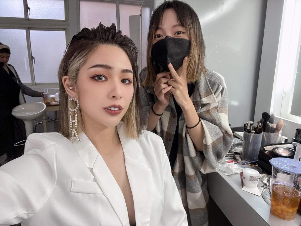 這張照片是一名女子在化妝鏡前,戴著珍珠耳環,穿著白色外套,側臉微笑,另一名女子則站在她身後,戴著口罩,身穿格紋外套,似乎正在替她化妝。