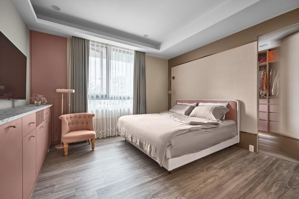這張「臥室」的照片是一張大床,床頭板是淺色,床鋪上鋪著灰色的床單,床邊有一個粉色的單人沙發,房間的牆壁上有一扇門,門後面是衣櫃,房間的地板是木地板。