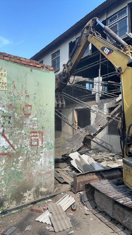 1) 照片顯示一輛黃色挖土機正在拆除一棟建築物,建築物外牆已經被拆除,可以看到建築物的內部,地上散落著瓦礫