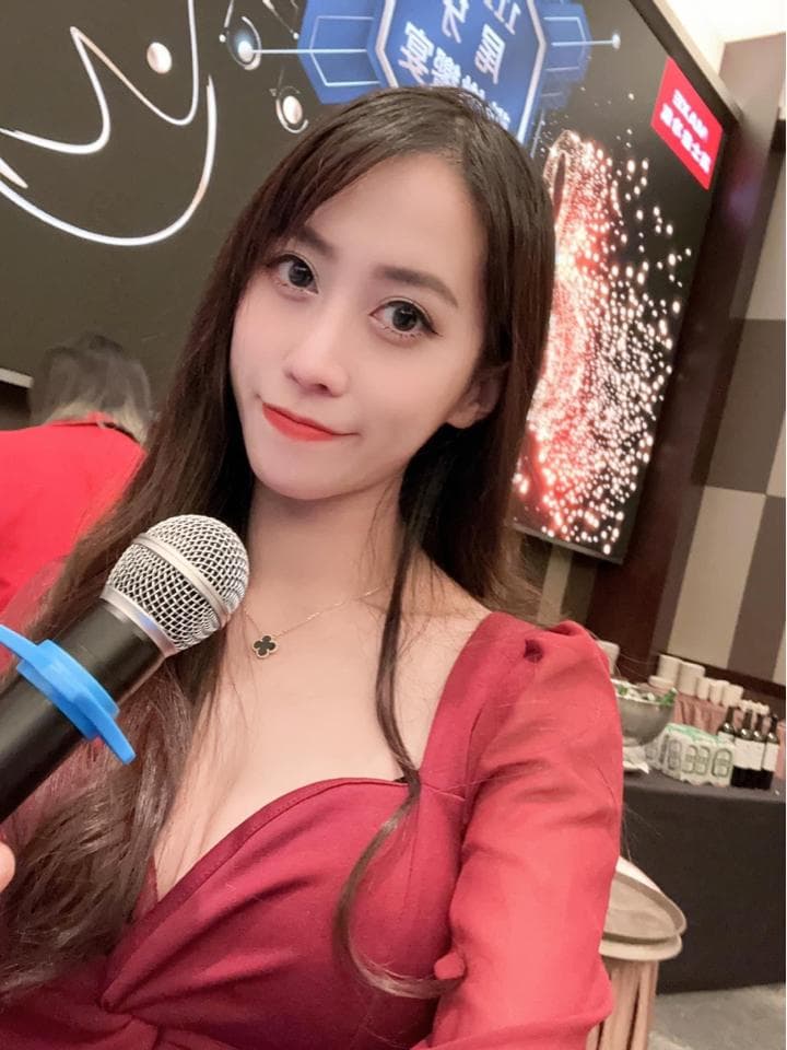 這張照片是一位穿著紅色洋裝的女性,她拿著麥克風,背景是模糊的燈光和一個大螢幕。