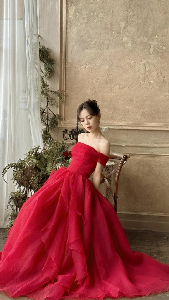 這張「一位女子身穿紅色禮服坐在椅子上」的照片是,她穿著一件紅色蓬裙禮服,露出香肩,坐在一把木頭椅子上,背景是牆壁和窗簾。
