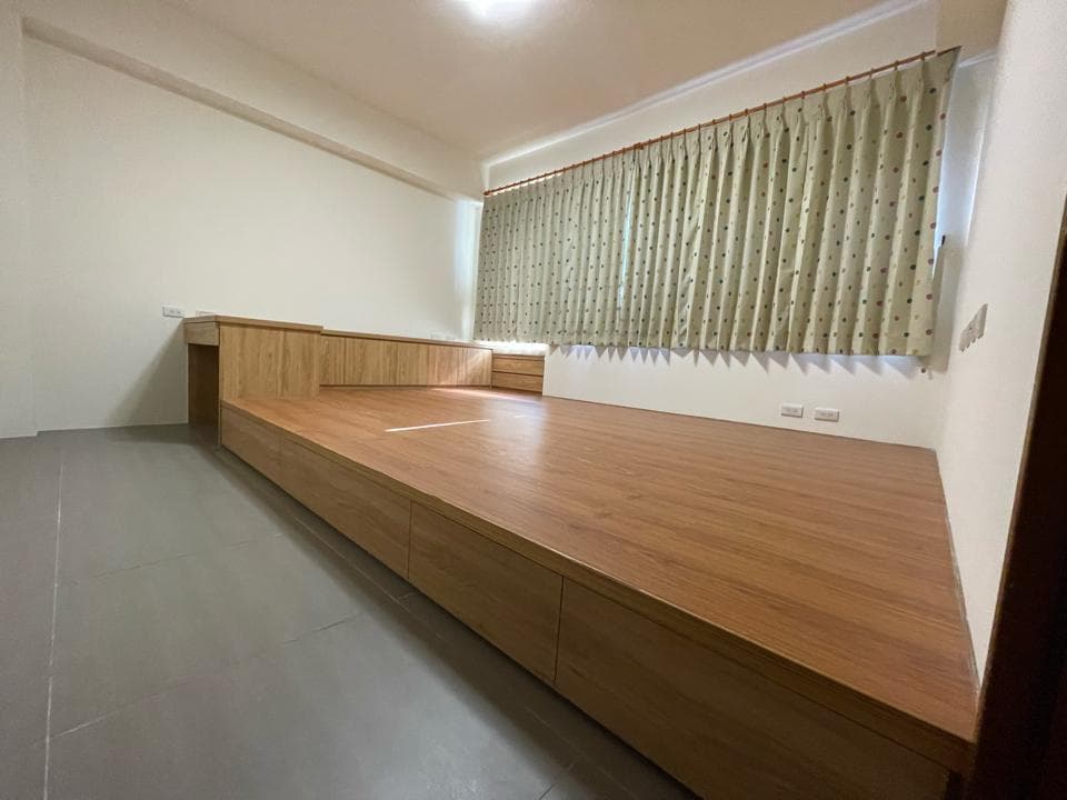 一张照片展示了一个木制平台床,它位于一间房间内,墙壁是白色的,房间里只有一扇窗户,窗户上装有米色的百叶窗帘,房间地板上铺着灰色瓷砖