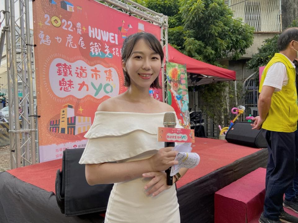 這張照片是一位穿著白色洋裝的女性,她拿著麥克風,背景是一塊紅色布條,上面寫著“歡遊市集 感恩 yo”。