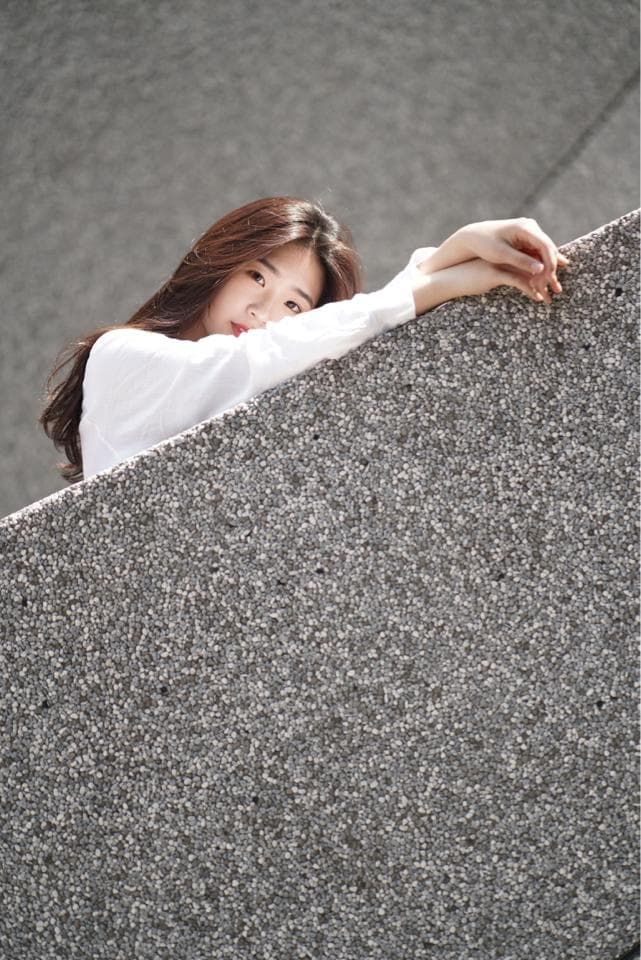 這張「一位女生靠在水泥牆上的」的照片是一名女生穿著白色襯衫,靠在水泥牆上,她的長髮散落在肩膀上,眼神望向鏡頭。