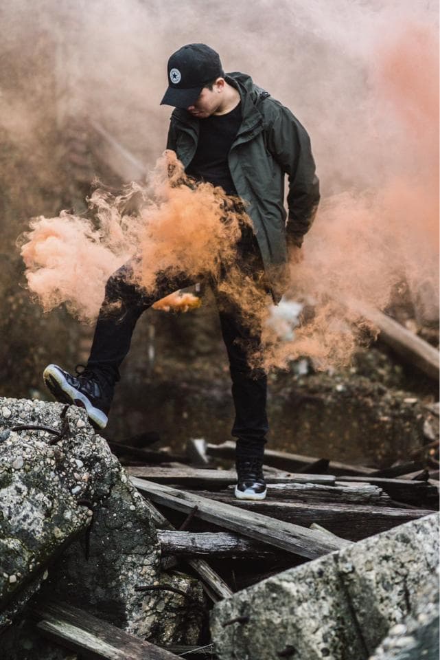 這張「一個戴著帽子的人在煙霧中行走」的照片是,一個戴著黑色帽子的男子站在廢墟中,他身旁環繞著橙色的煙霧,男子身穿黑色外套,腳踩黑色鞋子。