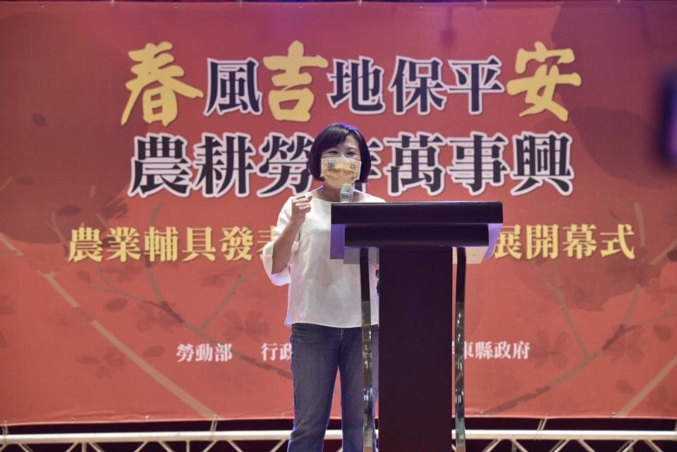 這張「女性站在講臺上演講」的照片,背景是紅色的布幕,上面寫著「春風故地保平安 農耕務實傳承」以及「農業界輔導說明儀式」。