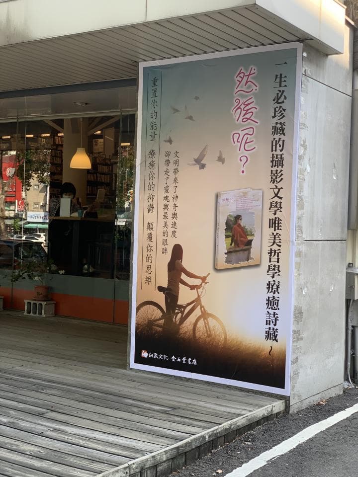 1) 這張照片中顯示了書店外牆的廣告看板,廣告看板上有圖案及文字,看板右側下方有一輛自行車,自行車後方有一位女性的剪影,看板右上角有一個人物的剪影以及幾隻鳥的剪影