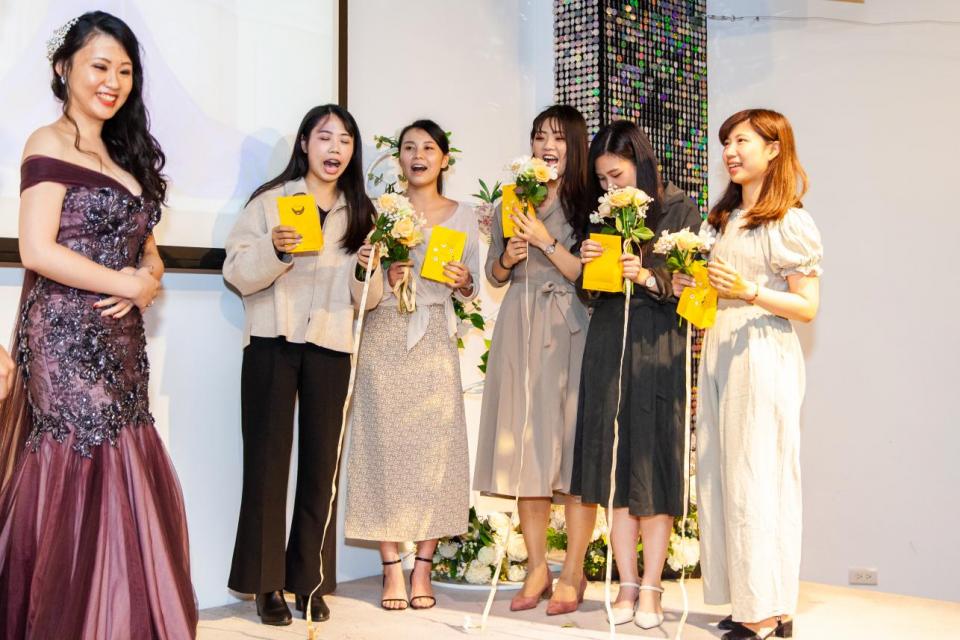 這張「一群女性在聚會中舉著禮物」的照片是,一位穿著紫色禮服的女性站在最前面,其他四位女性手持黃色禮盒,並配以花束,站在她身後。她們似乎都在慶祝一個特別的場合。