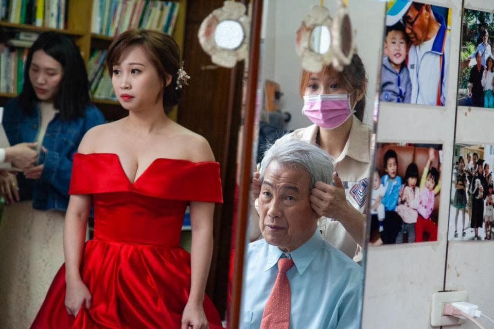 這張照片是 一個婚禮現場,新娘穿著紅色的婚紗,新郎坐在鏡子前準備,旁邊有一位女性幫忙整理新郎的頭髮。