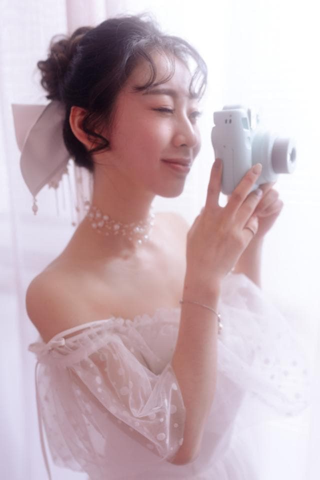 這張照片是一個年輕女子穿著白色蕾絲洋裝,她拿著一臺藍色的相機,眼睛望向鏡頭。