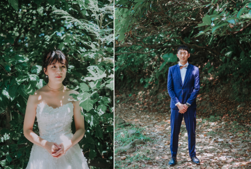 這張照片是一對穿著婚紗禮服的新人站在樹林裡。新郎穿著藍色西裝,打著領結,新娘穿著白色婚紗,他們都面向鏡頭,看起來很幸福。