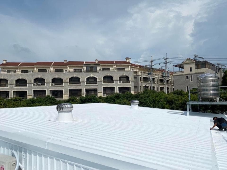 1) 照片顯示一棟建築物的屋頂,屋頂上有兩個白色圓形通風口,背景是一排白色建築物,周圍有樹木