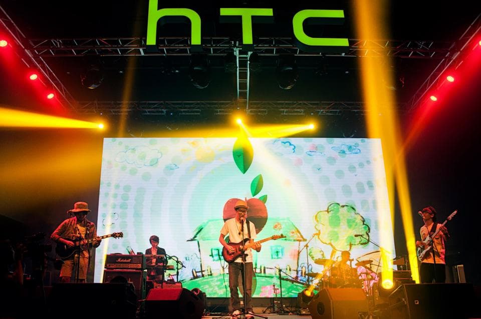 這張「HTC」的照片是一個音樂表演,舞臺上有一組樂隊正在演奏,他們身後有一個大的螢幕,螢幕上繪製著一些彩色圖案。