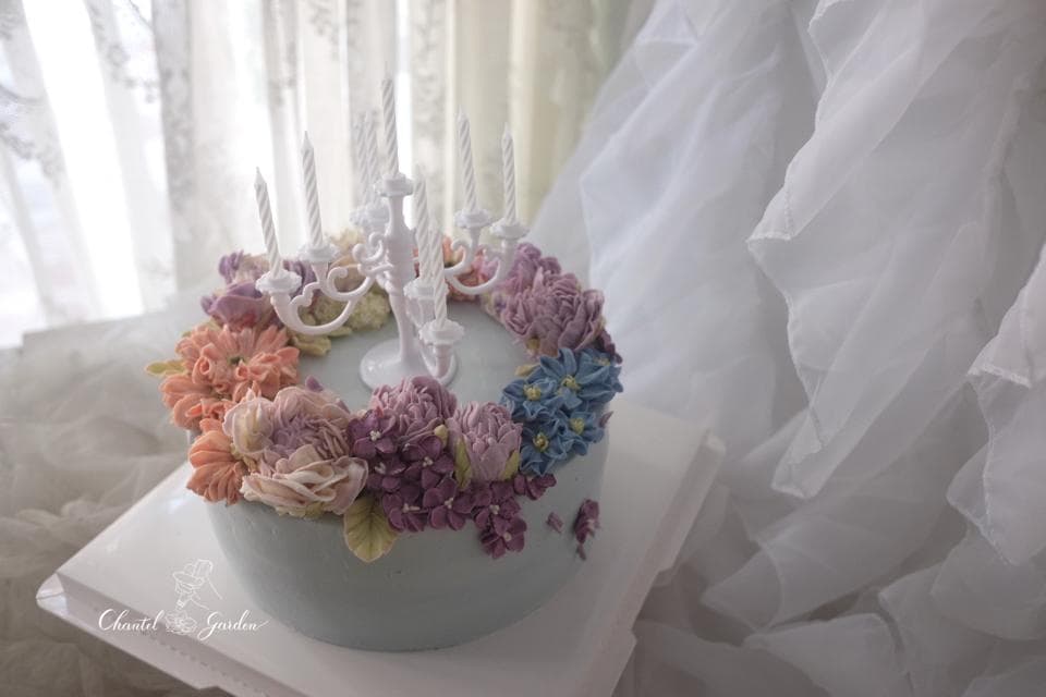 這張照片是一塊用奶油霜裝飾的蛋糕,周圍圍繞著淡粉紅色、淡紫色和藍色的花朵。蛋糕的中央是一個白色燭臺,上面有幾支蠟燭。蛋糕的顏色和設計非常優雅。