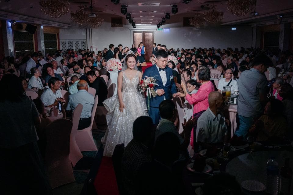 這張照片是婚禮的盛況,一對新人正接受賓客的祝福,新郎穿著黑色西裝,新娘穿著白色婚紗,他們手持花束,笑容滿面,現場賓客也熱烈鼓掌,洋溢著幸福的氛圍。