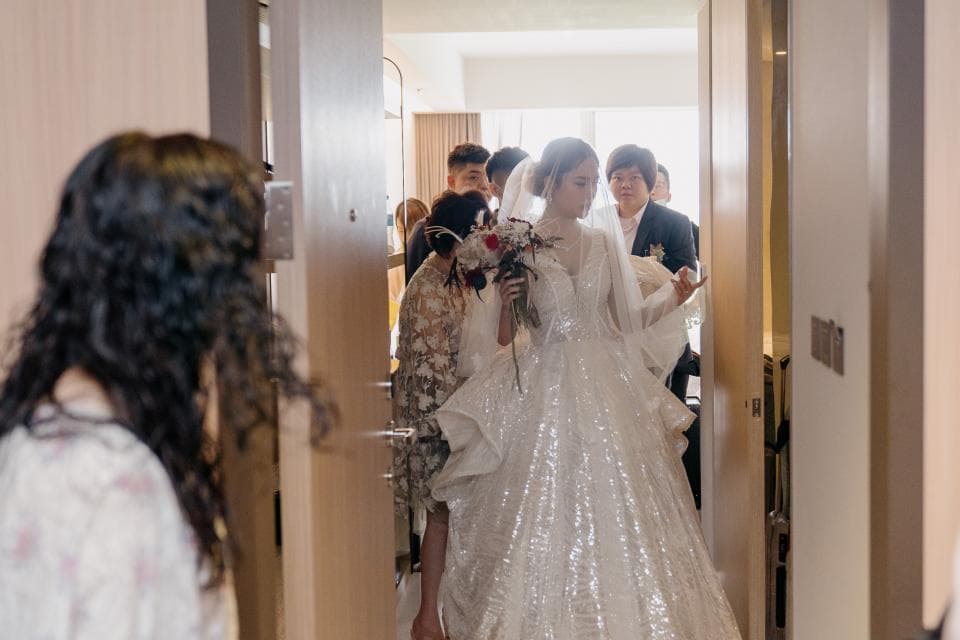 這張「婚禮」的照片是一個新娘從房間裡走出來,她穿著漂亮的婚紗,手裡拿著花束,旁邊有伴娘和家人陪同。