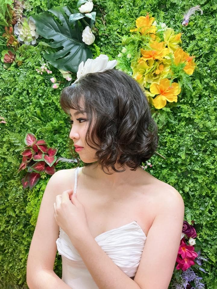 這張照片是一位留著短捲髮的女性,她戴著白色的花飾,穿著白色連衣裙,站在綠色的植物牆前,她低頭看著右手,呈現出害羞的模樣。