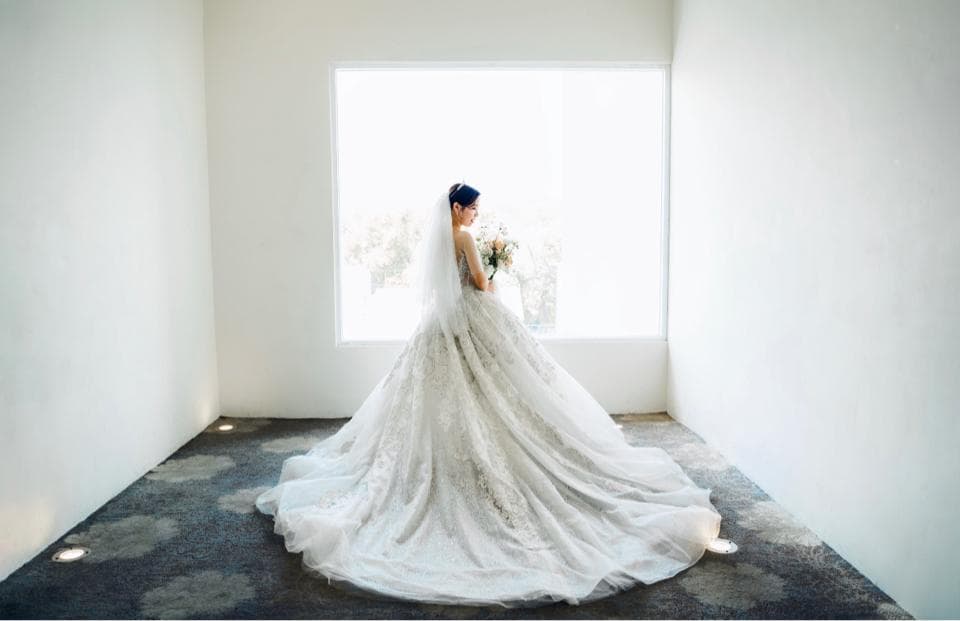 這張婚禮照片是一個穿著白色婚紗的新娘,她站在一個窗戶前,手拿著花束,面帶微笑。