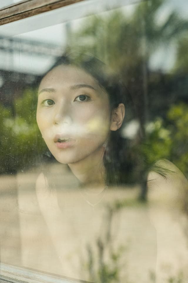 這張照片是一名女性從窗戶向外看的模糊身影。她的臉部輪廓、眼睛和嘴巴都清晰可見,但她的背景卻被模糊處理,營造出一種神秘的氛圍。透過窗戶的玻璃,可以隱約看到她身後的綠色植物,暗示著窗戶外的自然景觀。