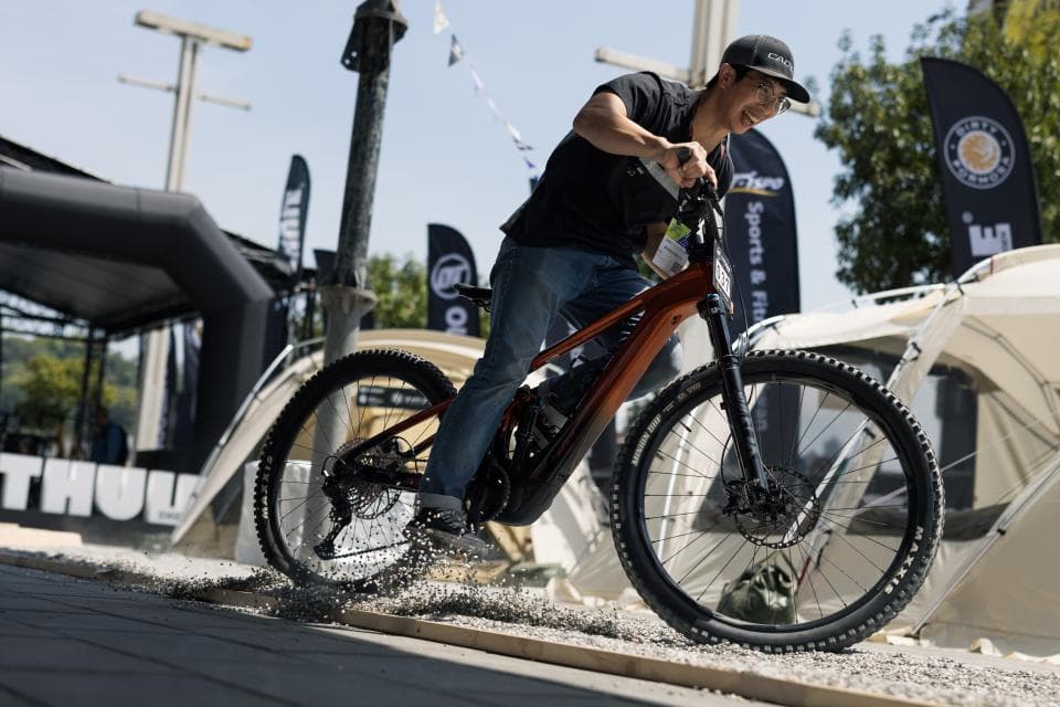 這張「男子騎著山地自行車」的照片是,他穿著短袖和長褲,戴著帽子,正騎著一輛橘色的山地自行車,他身處在戶外,背景是一些棚子。