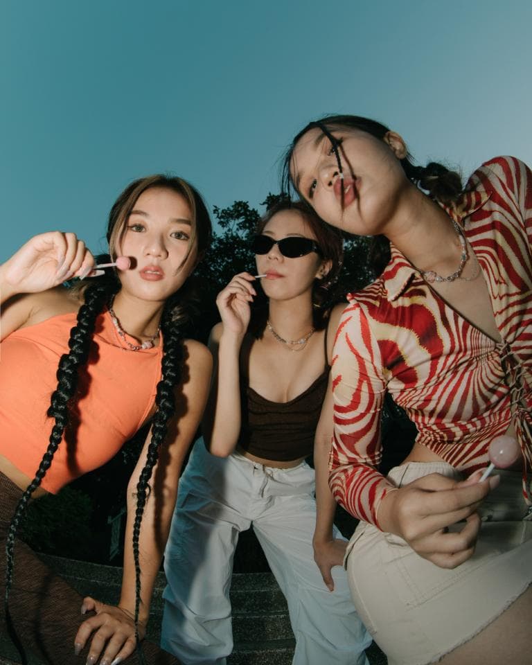 這張「三個女生在戶外玩耍」的照片是三個女孩在夕陽下,戴著太陽眼鏡,穿著不同的服裝,拿著棒棒糖,一起拍照。