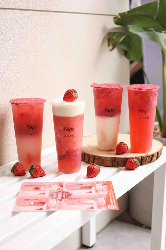 這張「草莓飲品」的照片是四杯草莓飲品放在白色木桌上,周圍擺放著草莓,還有一張印有草莓圖案的菜單。