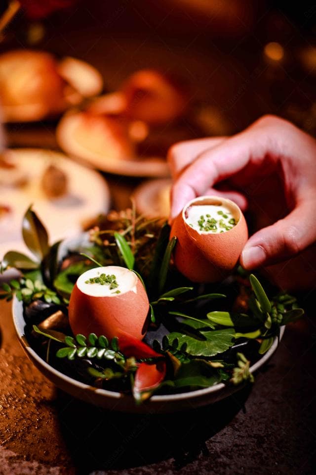 這張「蛋殼甜點」的照片是兩顆蛋殼裝的甜點,上面撒了綠色的香草,放在綠色的植物中,背景是昏暗的燈光。