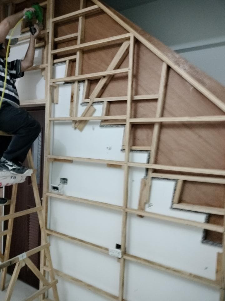 這張照片是一名工人站在梯子上,正在用電鑽固定牆上的木條。工人穿着黑色的衣服,手裡拿着一個綠色的電鑽,正對着牆壁操作。牆上已經安裝了一些木條,並形成了一个網格狀結構,看起來像是正在進行装修工程。