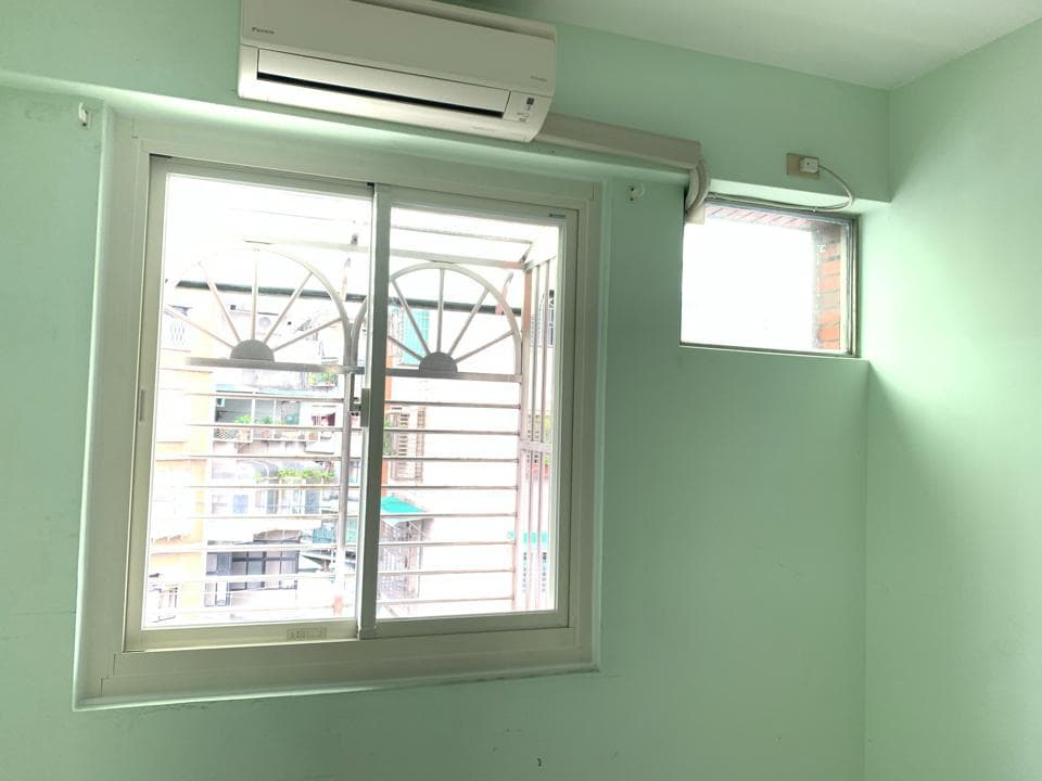1) 照片顯示一間房間內部,房間牆壁是淺綠色的,房間內有一台冷氣、一個大窗戶,窗戶外面是其他建築物,以及一個小窗戶