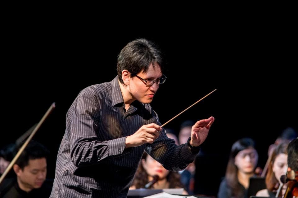 這張照片是一個正在指揮樂團的指揮家。他穿著黑色襯衫,手持指揮棒,專注地注視著樂團。