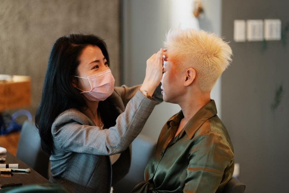 這張照片是一個女人正在為另一個女人化妝,她們在一個工作室裡,攝影師在拍她們。