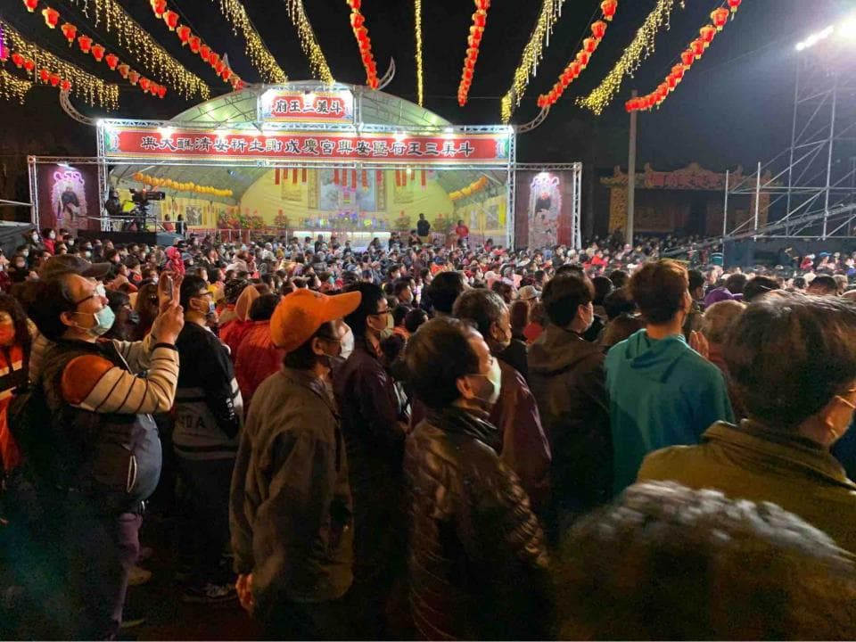 這張照片是人群聚集在一個戶外活動中,人們穿著不同的服裝,背景是彩色的燈光裝飾和一個舞臺,看起來非常熱鬧。