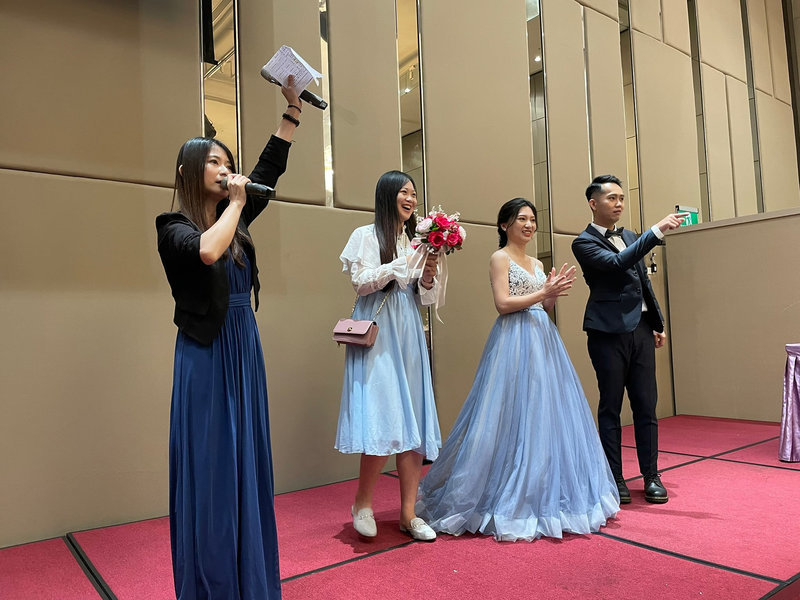 這張「婚禮」的照片是一個婚禮上的遊戲環節,一對新人和兩位主持人站在臺上。 新娘穿著淡藍色的禮服,新郎穿著西裝,主持人正在講話,新娘則手持捧花。