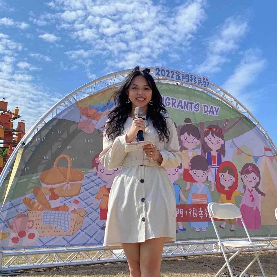 這張照片是一個女生站在一個大型佈景前,佈景上有「移民節」的字樣。女生穿著白色洋裝,手拿麥克風,笑容滿面。