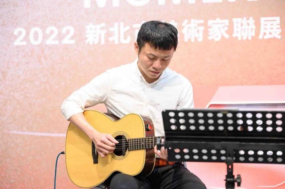 這張照片是一個年輕男子正在彈吉他的畫面。他穿著白襯衫,專注地演奏著吉他,手上的動作流暢自然,臉上帶著滿足的笑容。他面前放著一個樂譜架,暗示著他正在演奏一首特定的歌曲。