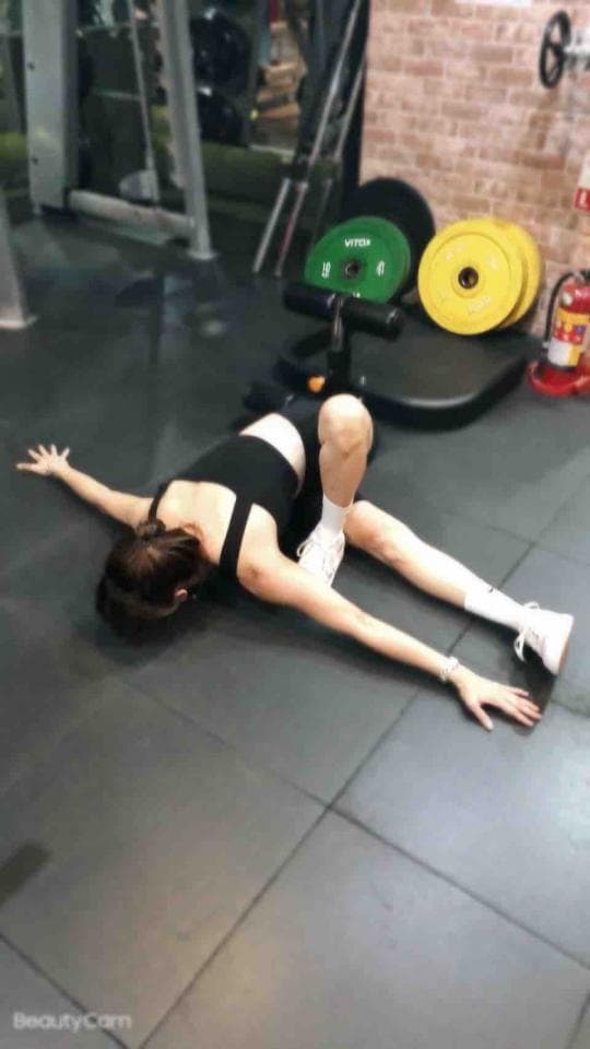 這張照片是一個女人在健身房地板上做伸展運動。她穿著黑色運動背心和白色運動鞋,雙手伸直,身體呈弓形,看起來很柔軟。