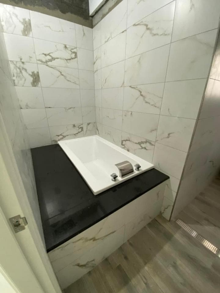 1) 照片顯示了一個裝修中的浴室,有一個嵌入式的浴缸,牆壁和地板都貼了白色大理石瓷磚,浴缸上放著一個黑色的平台,平台上有一個黑色的水龍頭