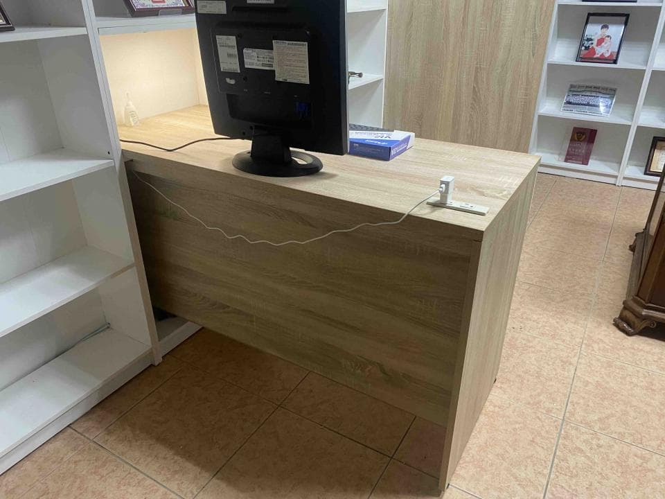 這張照片是一張木製書桌,桌面為木紋色,桌面上有電腦、線路、插座等。桌面下方是白色的櫃子。