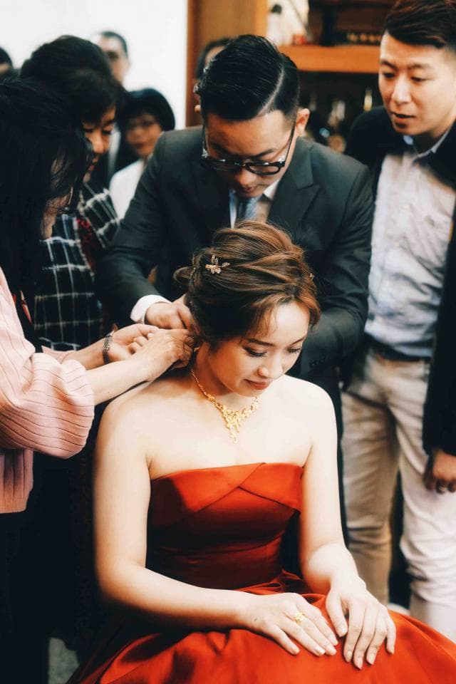 這張照片是一個穿著紅色禮服的新娘,她正坐在椅子上,讓一位穿著西裝的男人幫她整理頭髮。