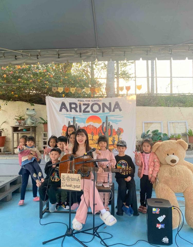 這張「一群小朋友在戶外聚會唱歌」的照片是,一位女生在舞臺上彈吉他唱歌,周圍圍繞著一群小朋友,還有一隻泰迪熊。