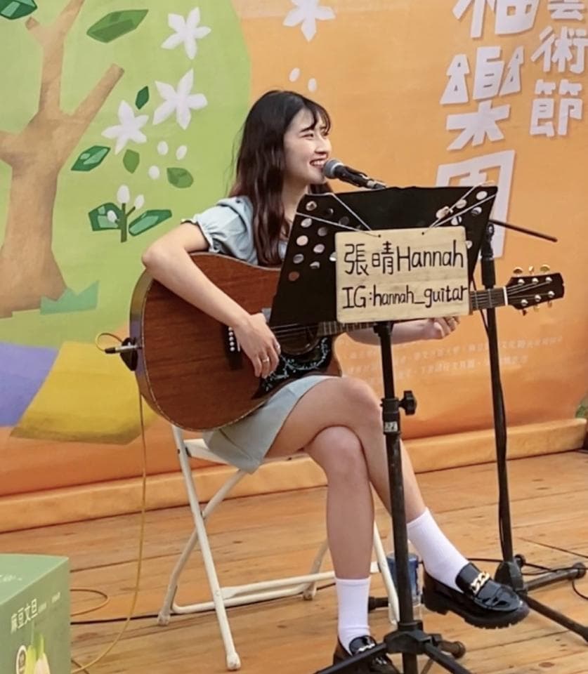 這張「一位年輕女孩正在戶外彈吉他的照片」是,她穿著藍色上衣,坐在一把摺疊椅上,並專注地彈著吉他。