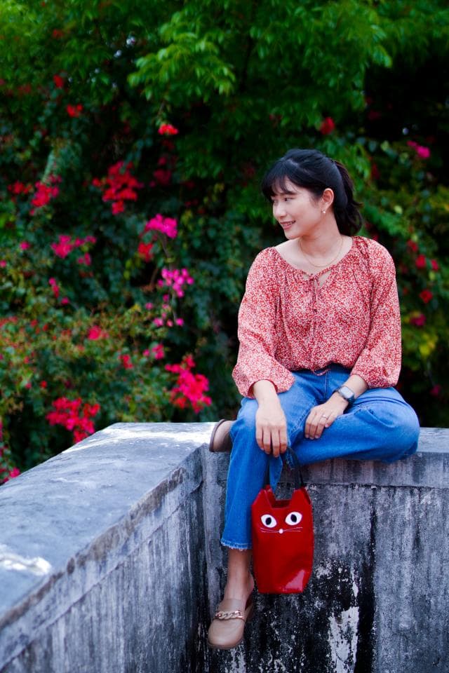 這張照片是一名女子坐在石牆上,她穿著紅色的上衣和藍色的牛仔褲,手裡拿著一個紅色的貓咪形狀的小包,看起來很可愛。