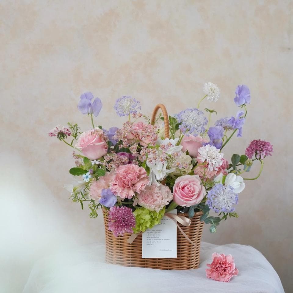 這張照片是一個裝滿粉紅色和紫色花朵的藤製籃子。籃子裡的花朵包括康乃馨、玫瑰、滿天星和其他幾種花卉,它們被排列成一個美麗的裝飾。