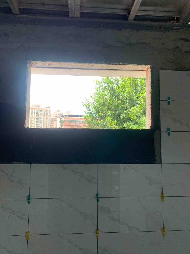 這張照片是正在裝修的浴室,牆上貼著白色磁磚,窗戶正對著綠色樹木,窗外可見建築物的屋頂。