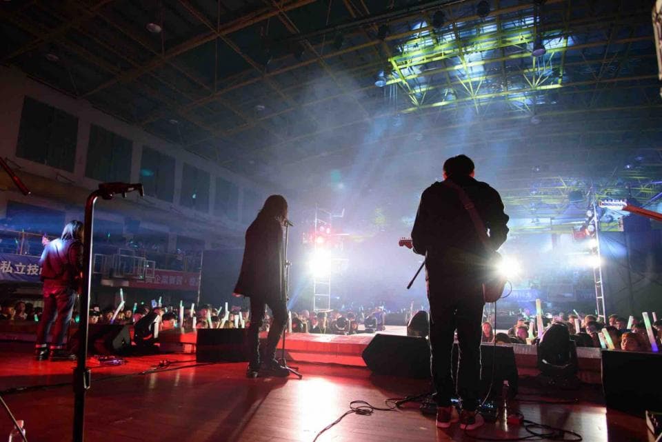 這張照片是一個音樂會現場,一個吉他手正在舞台上表演。他身穿黑色夾克,正在彈奏電吉他,前方有聚光燈照射。他的身影被灯光照亮,顯得十分帥氣。