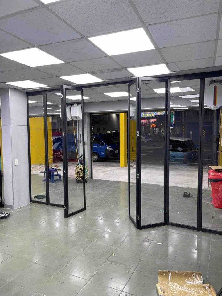 這張照片是一間現代化的空間,中間有一道黑色玻璃折疊門,門後方是另一個空間,空間明亮,並可看到一輛藍色的汽車停在門外。